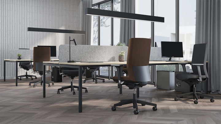 An office space with bench desks and ergonomic task chairs, privacy screens between the workstations, computers on the desks, and large windows with sheer curtains, in a room with herringbone pattern wood flooring.