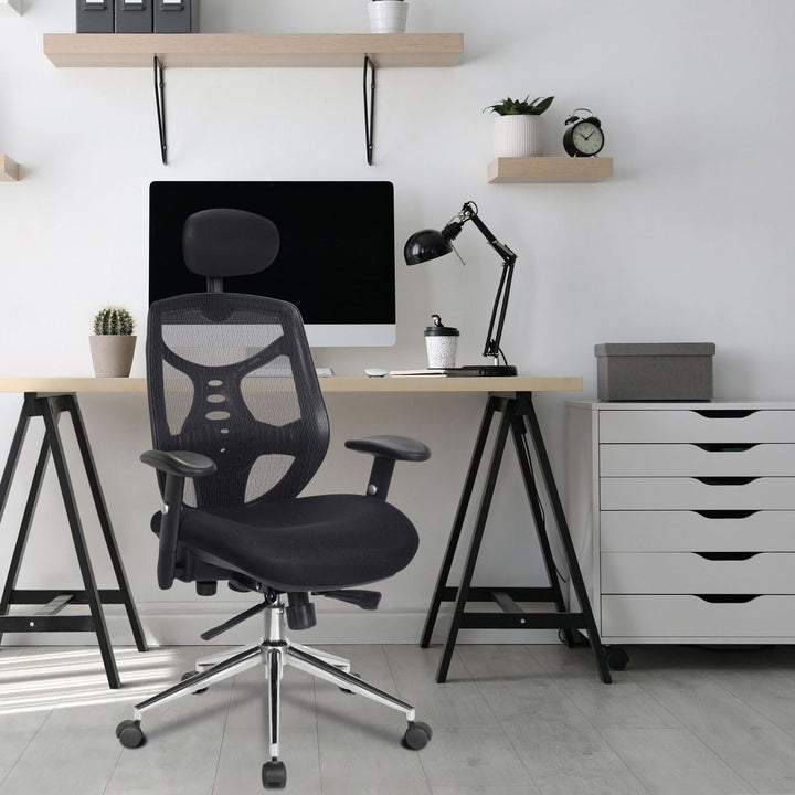 An ergonomic office chair with black mesh backrest and seat, adjustable armrests, and a five-wheeled chrome base sits in front of a desk with a computer monitor, a black desk lamp, and various office supplies.